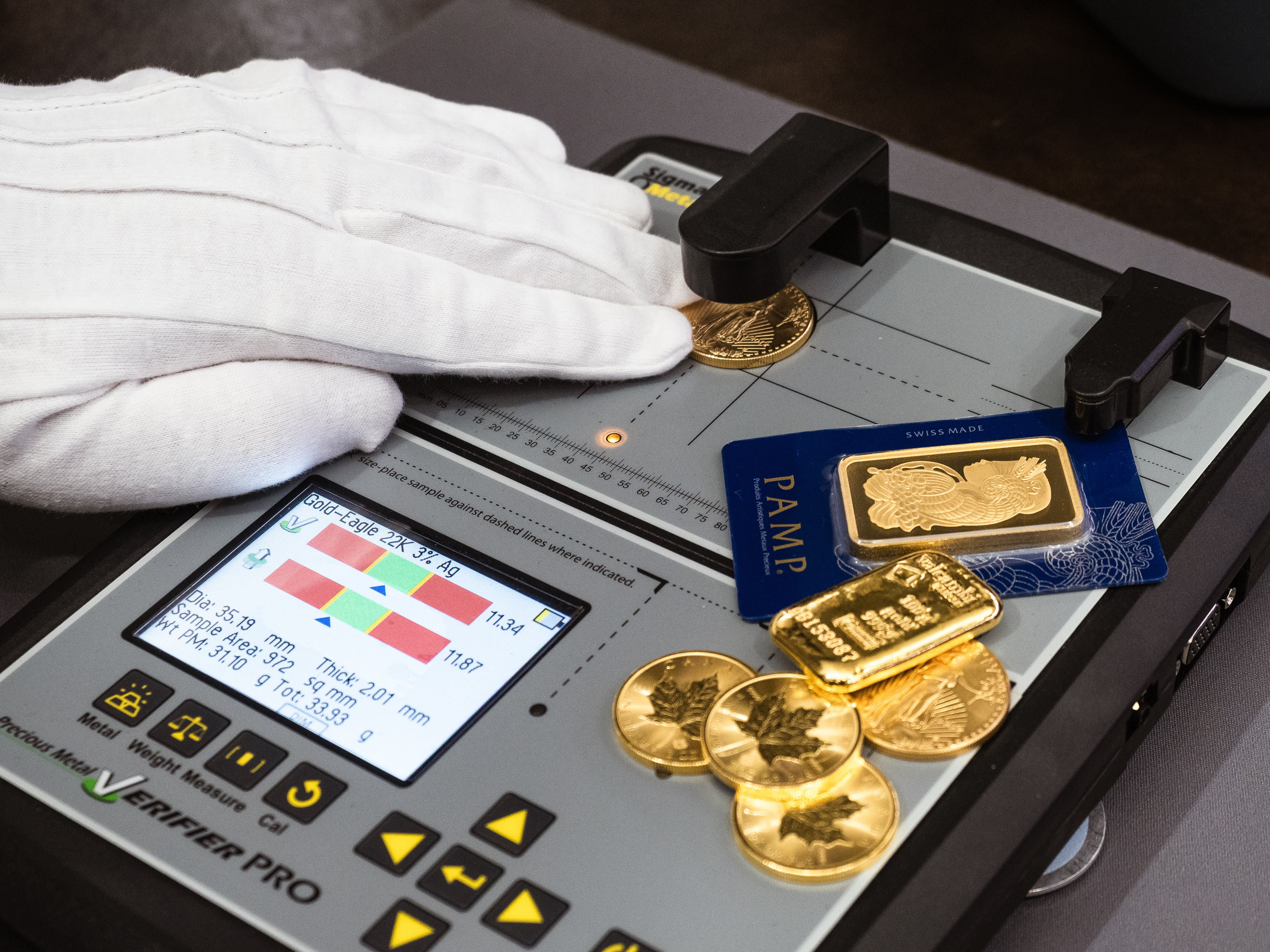 Gloved hand testing a gold coin on an electronic precious metal verifier, with PAMP and Valcambi gold bars and Maple Leaf coins beside the device