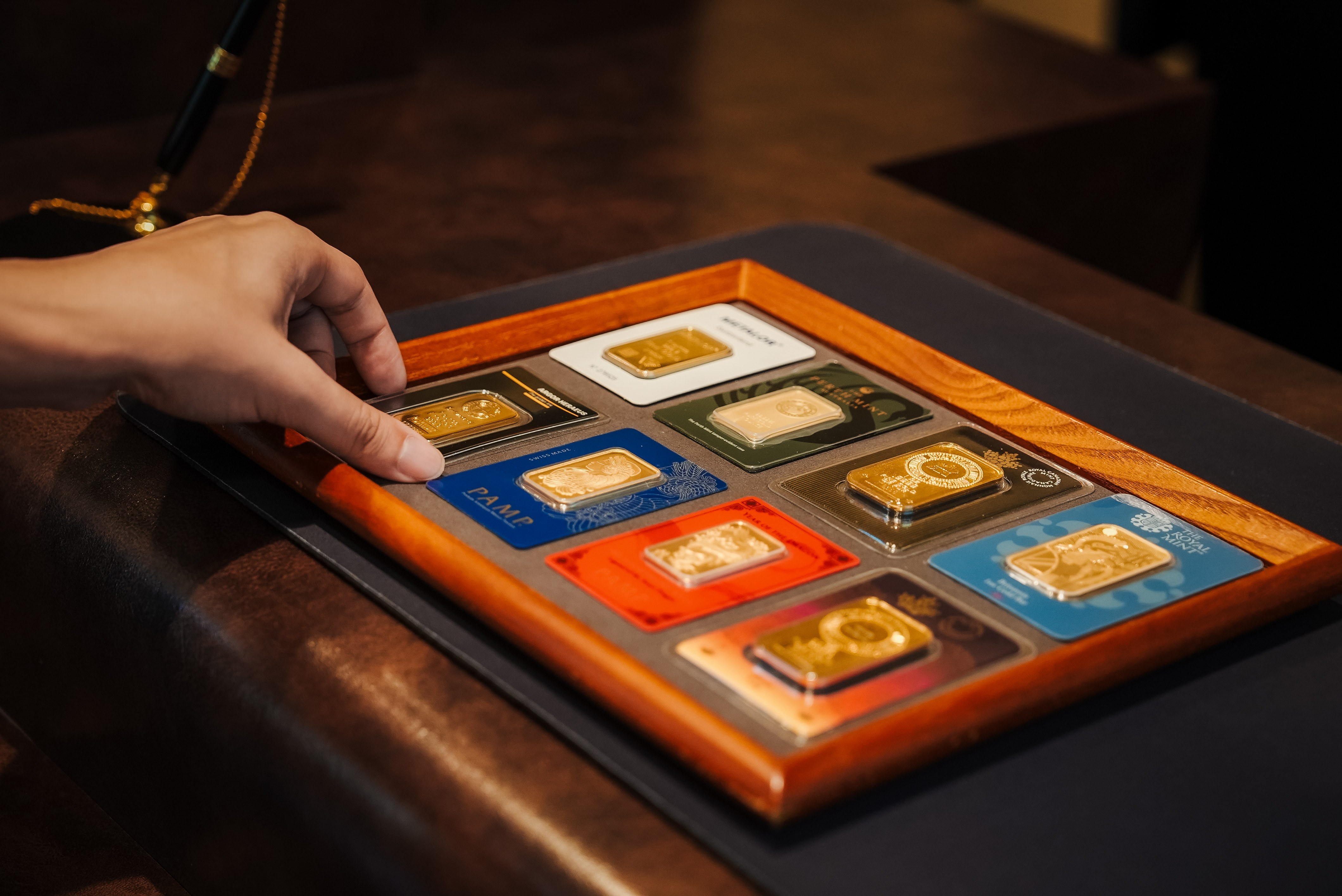  Hand selecting a minted gold bar from a display tray holding bars from PAMP, Perth Mint, Royal Mint, and other refiners