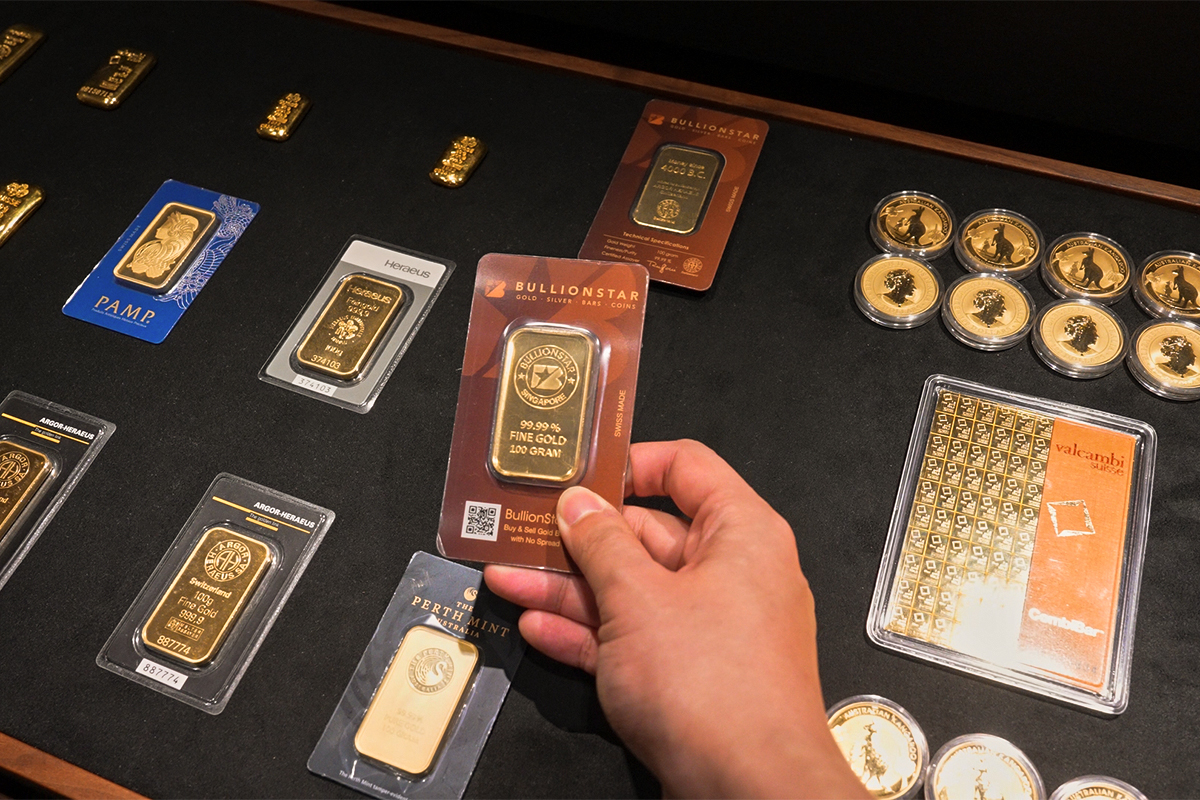 Hand holding a sealed BullionStar 100-gram fine gold bar above a display of bars from PAMP, Heraeus, and Perth Mint