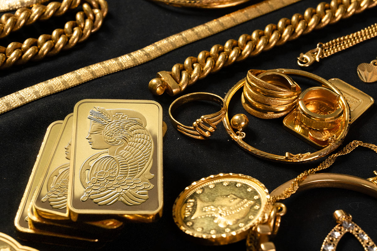 Second hand gold jewelry including chains, rings, and a bangle, arranged alongside gold bars stamped "Geneva" and a gold coin on a black surface