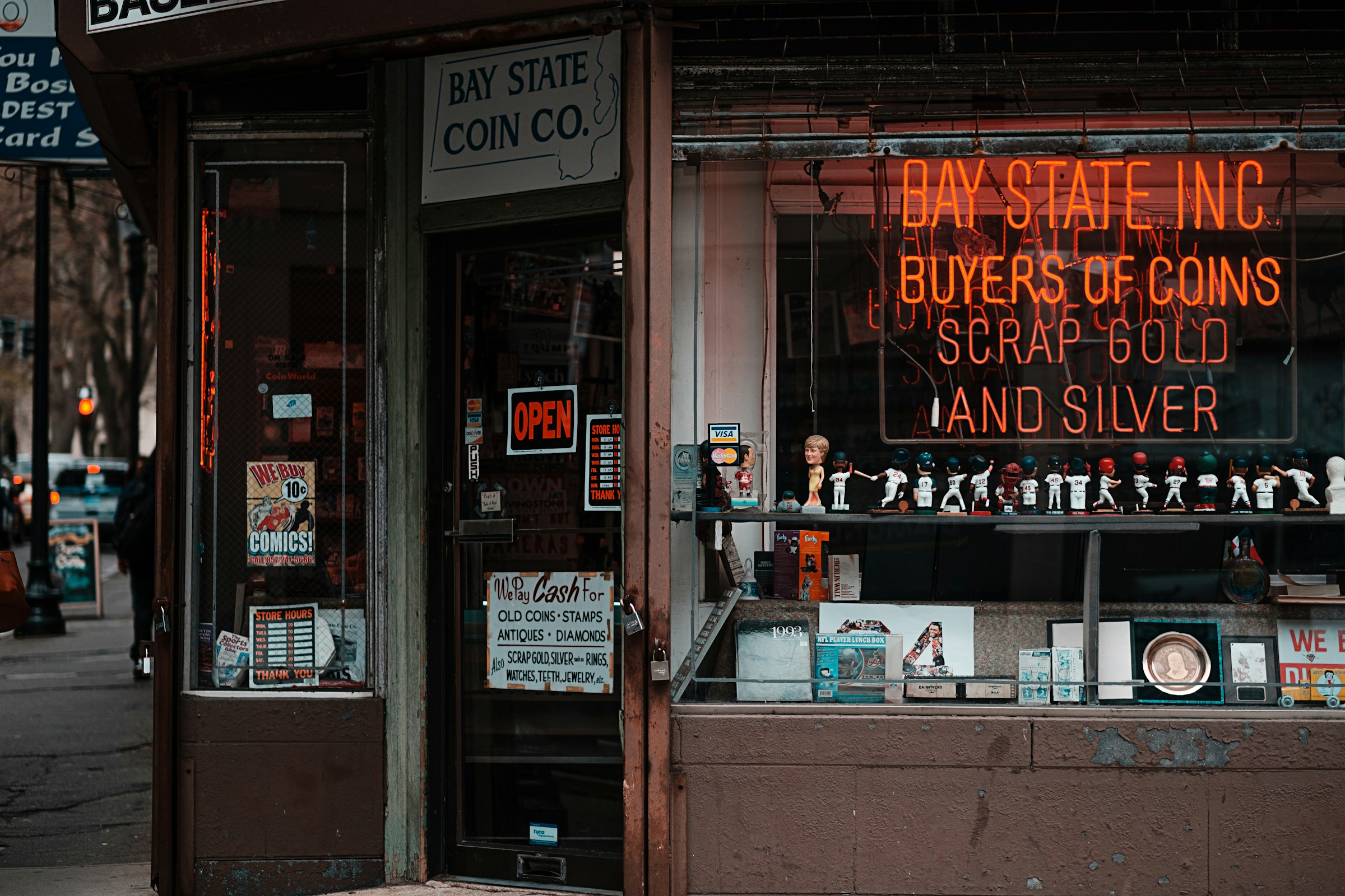 Pawn shop storefront at dusk with a neon sign reading "Bay State Inc, Buyers of Coins, Scrap Gold and Silver," alongside window displays of coins, comics, and collectibles