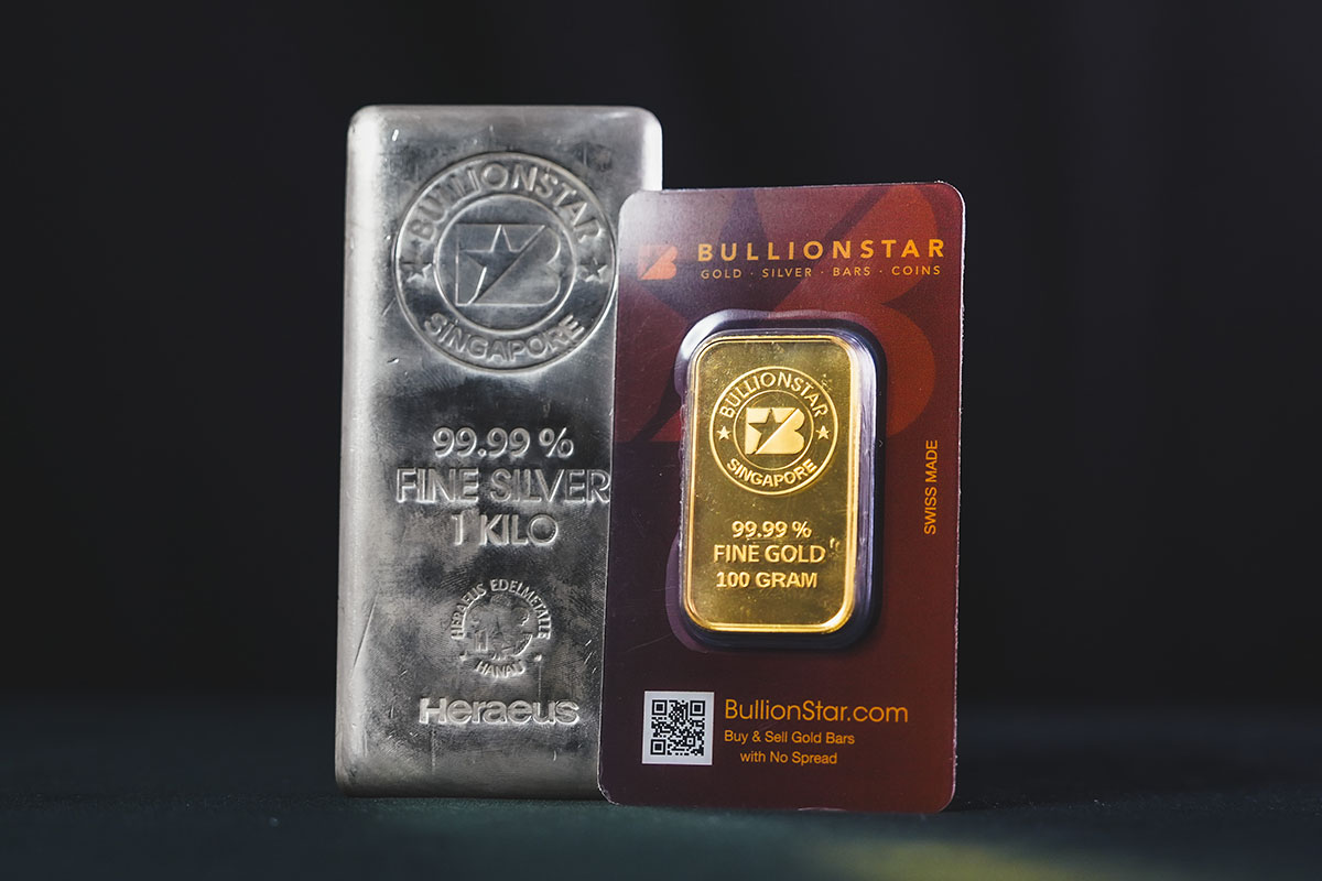 BullionStar 100-gram 99.99% fine gold bar in branded packaging alongside a BullionStar 1-kilo 99.99% fine silver bar