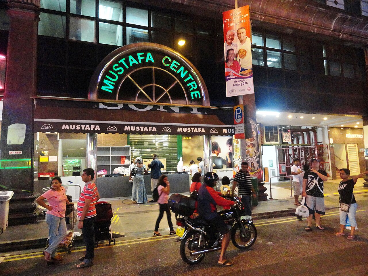 Mustafa Centre storefront at night on Syed Alwi Road in Little India, Singapore, with an illuminated sign and shoppers on the street outside