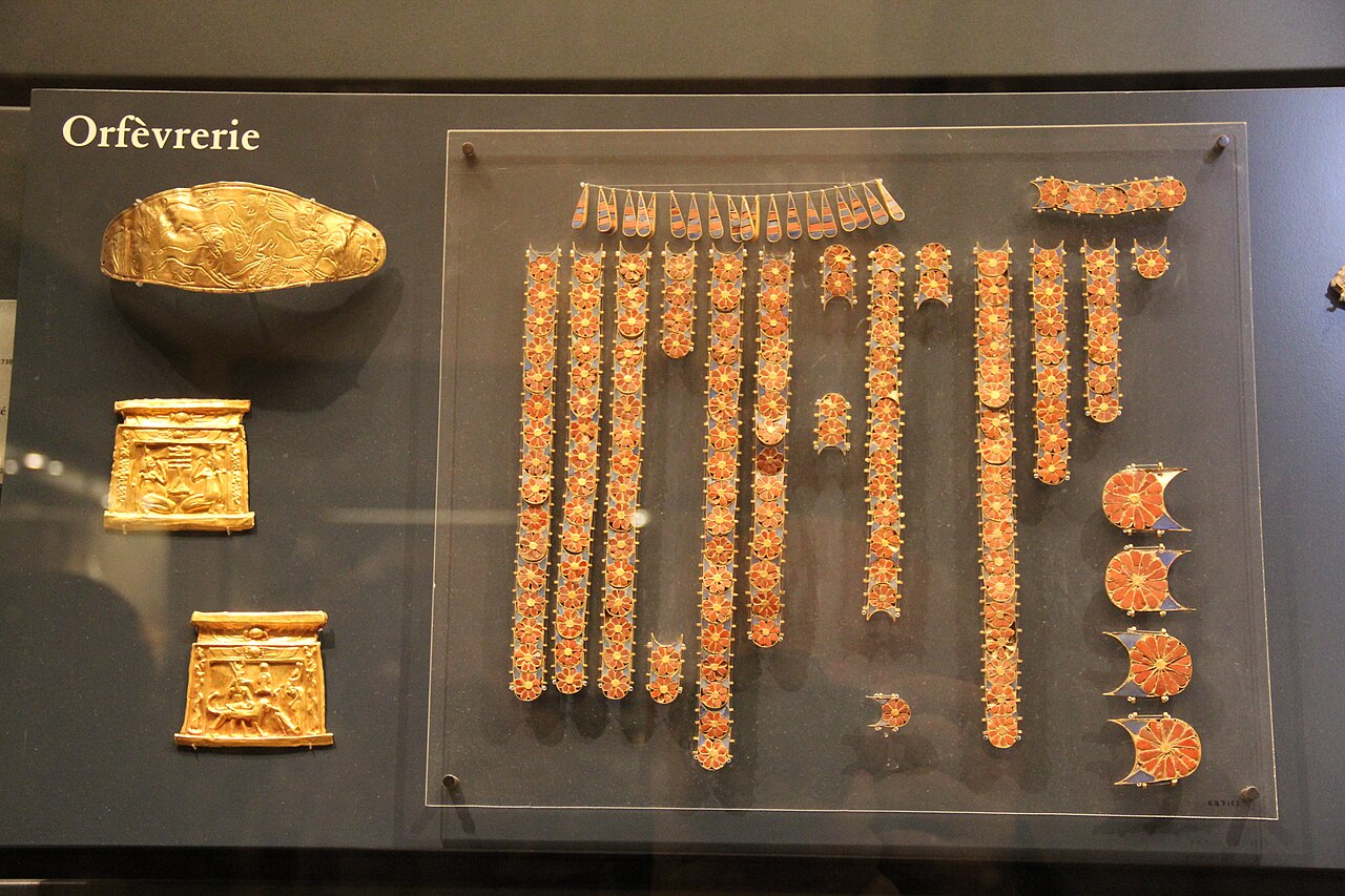Museum case labelled "Orfèvrerie" displaying ancient Egyptian goldwork, including three gold plaques worked in relief and rows of gold-mounted red and blue inlaid jewellery elements