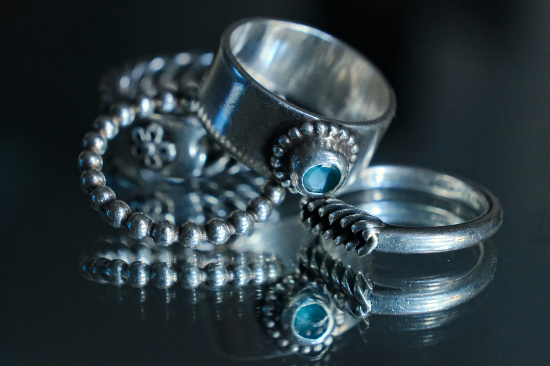 Three silver rings with oxidised detailing, including a beaded band, a wide plain band and a ring set with a blue stone, arranged on a reflective black surface