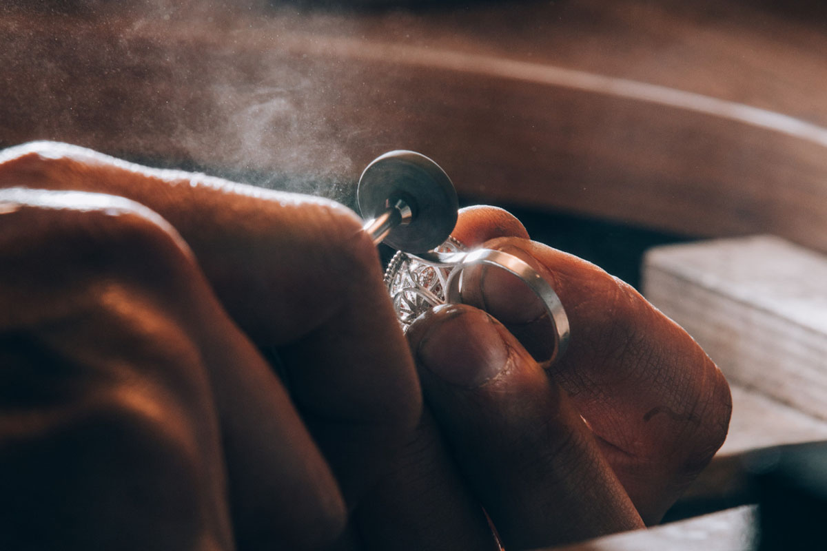 Close-up of a jeweller's hands holding a pale metal ring set with small stones against a rotary polishing wheel, with fine dust caught in the light